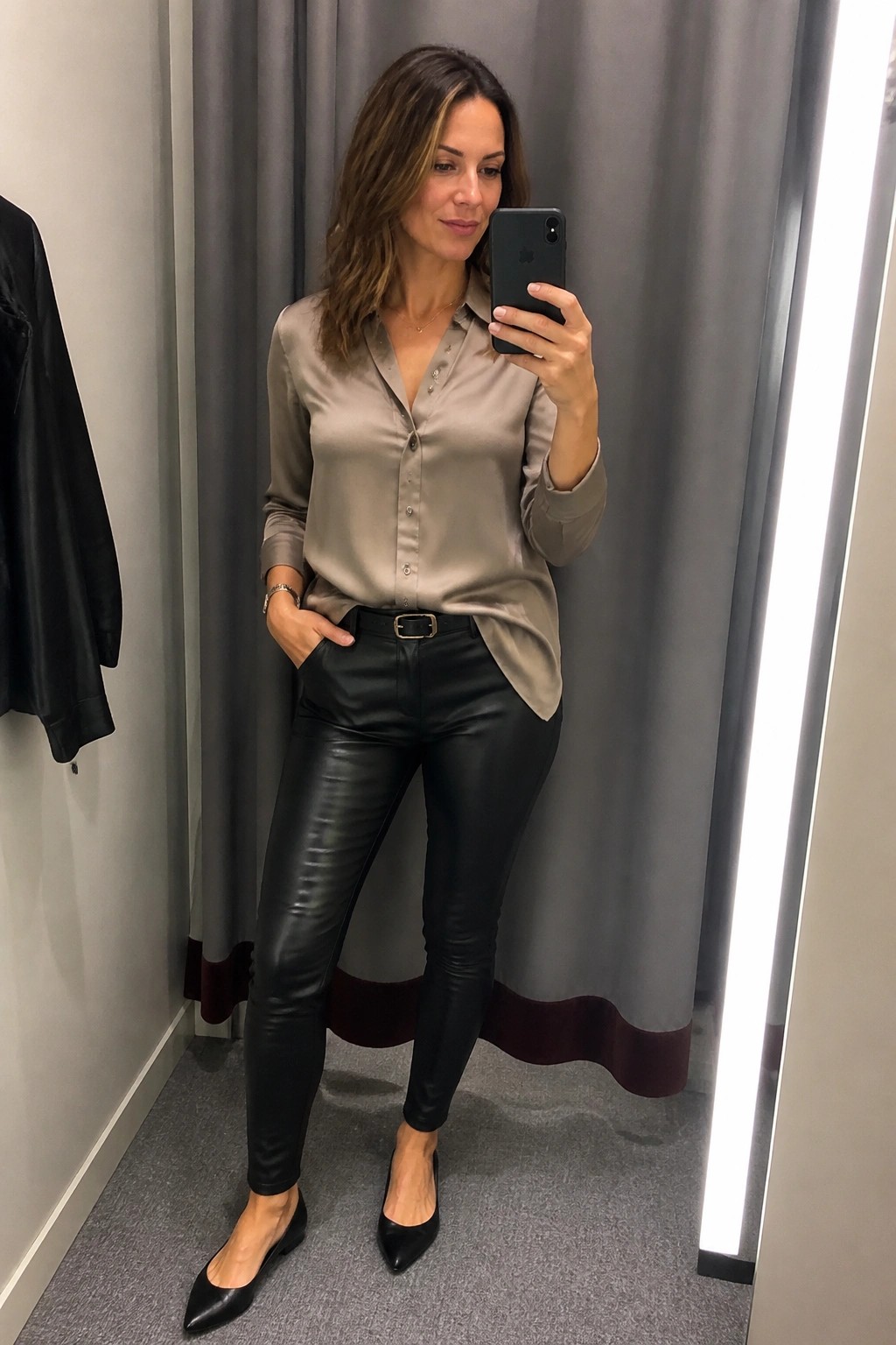 Woman in beige satin blouse and black leather pants taking mirror selfie in fitting room