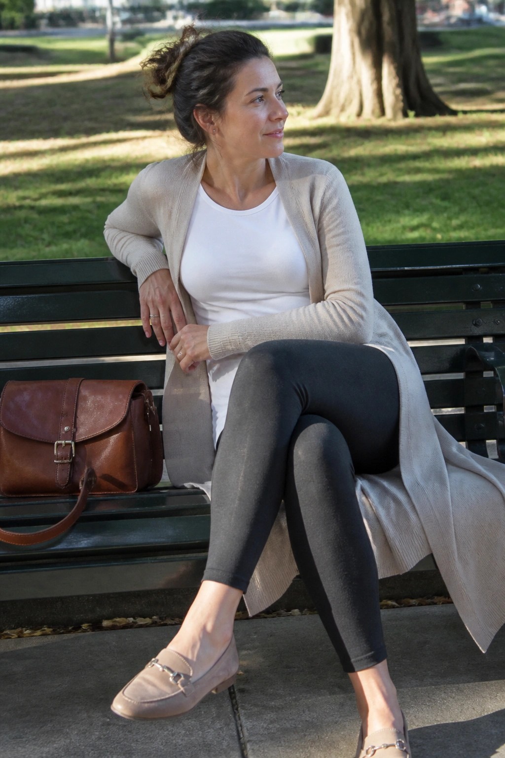 Woman in beige cardigan and black leggings on park bench with brown leather bag