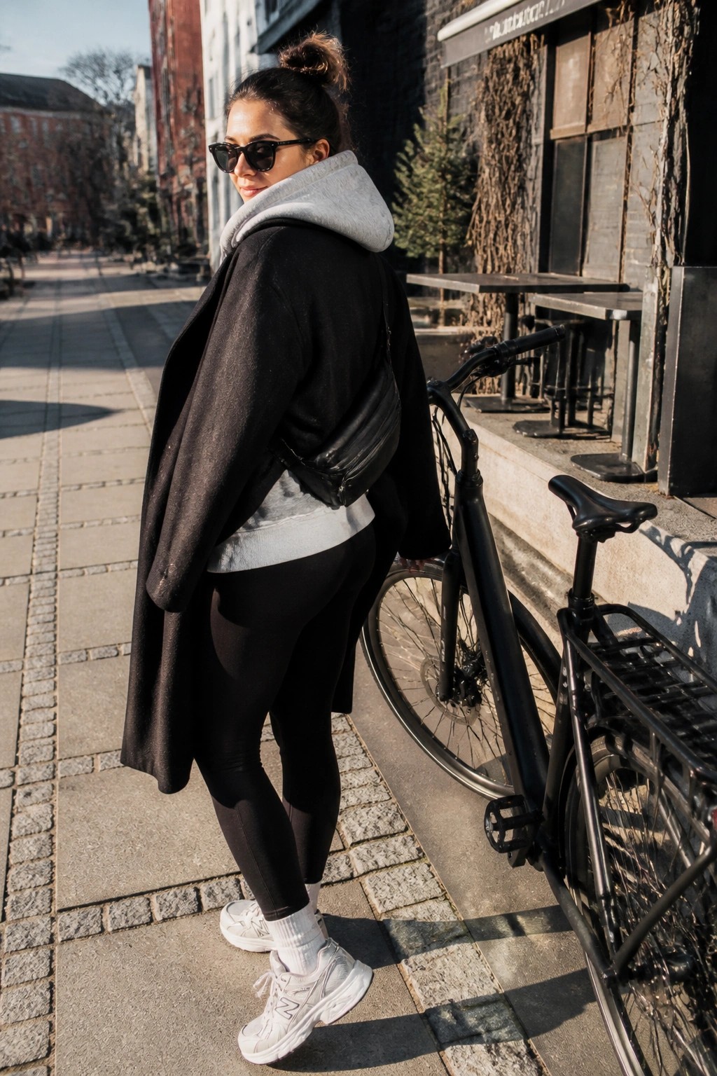 Woman in black coat and sunglasses beside bicycle on sunny city sidewalk