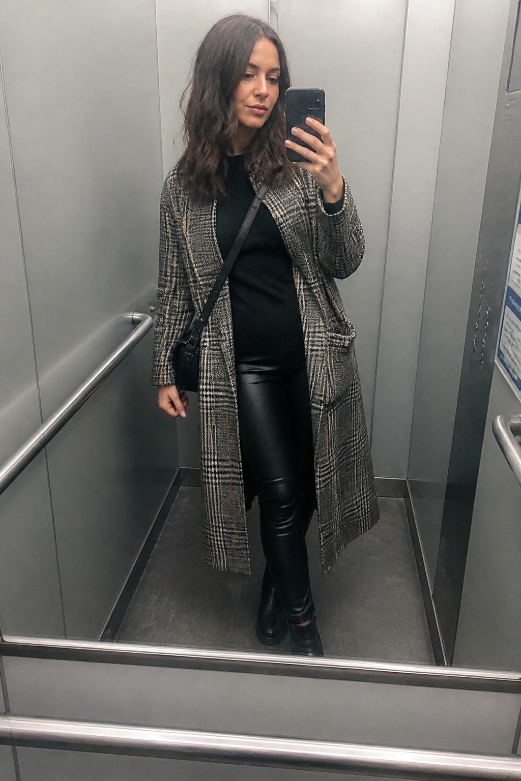 Woman in plaid coat and leather pants taking mirror selfie in elevator