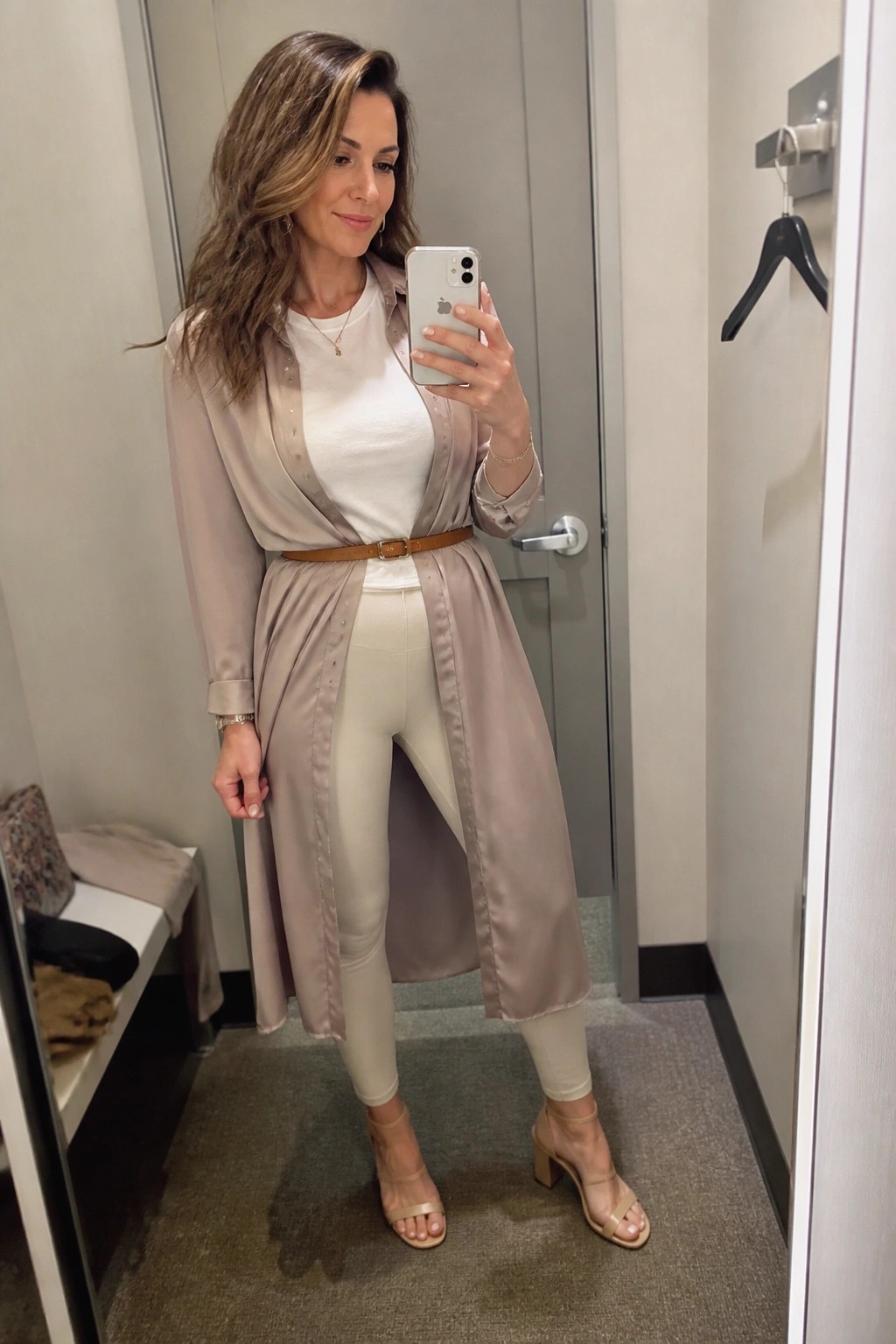 Woman in beige satin coat taking mirror selfie in fitting room with beige heels