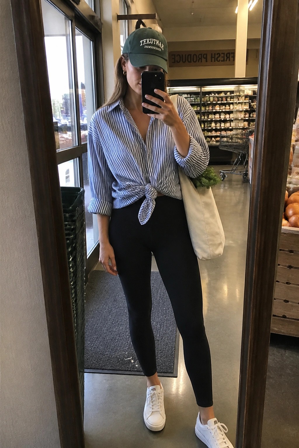 Woman in green cap, tied striped shirt, black leggings taking mirror selfie in grocery store.