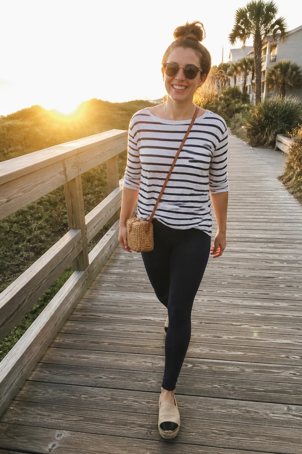 Woman in striped shirt and black leggings on sunset boardwalk with woven bag