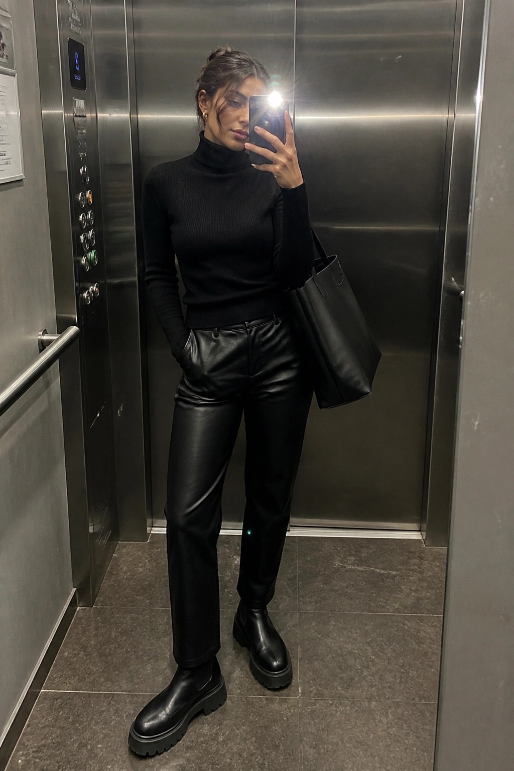 Woman in black turtleneck and leather pants taking mirror selfie in elevator.