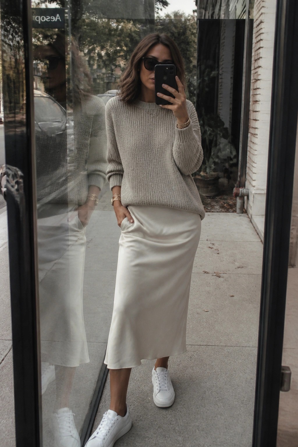 Woman in beige knit sweater and white satin skirt taking mirror selfie outdoors