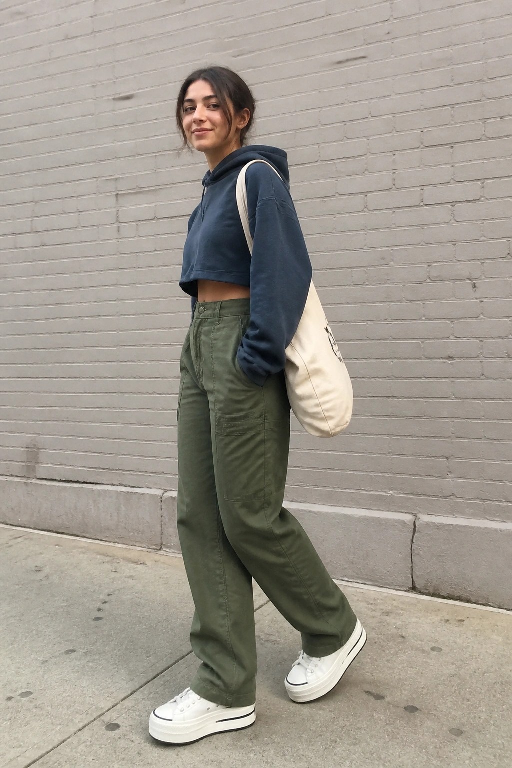 Woman in cropped navy hoodie and green cargo pants with tote bag by brick wall