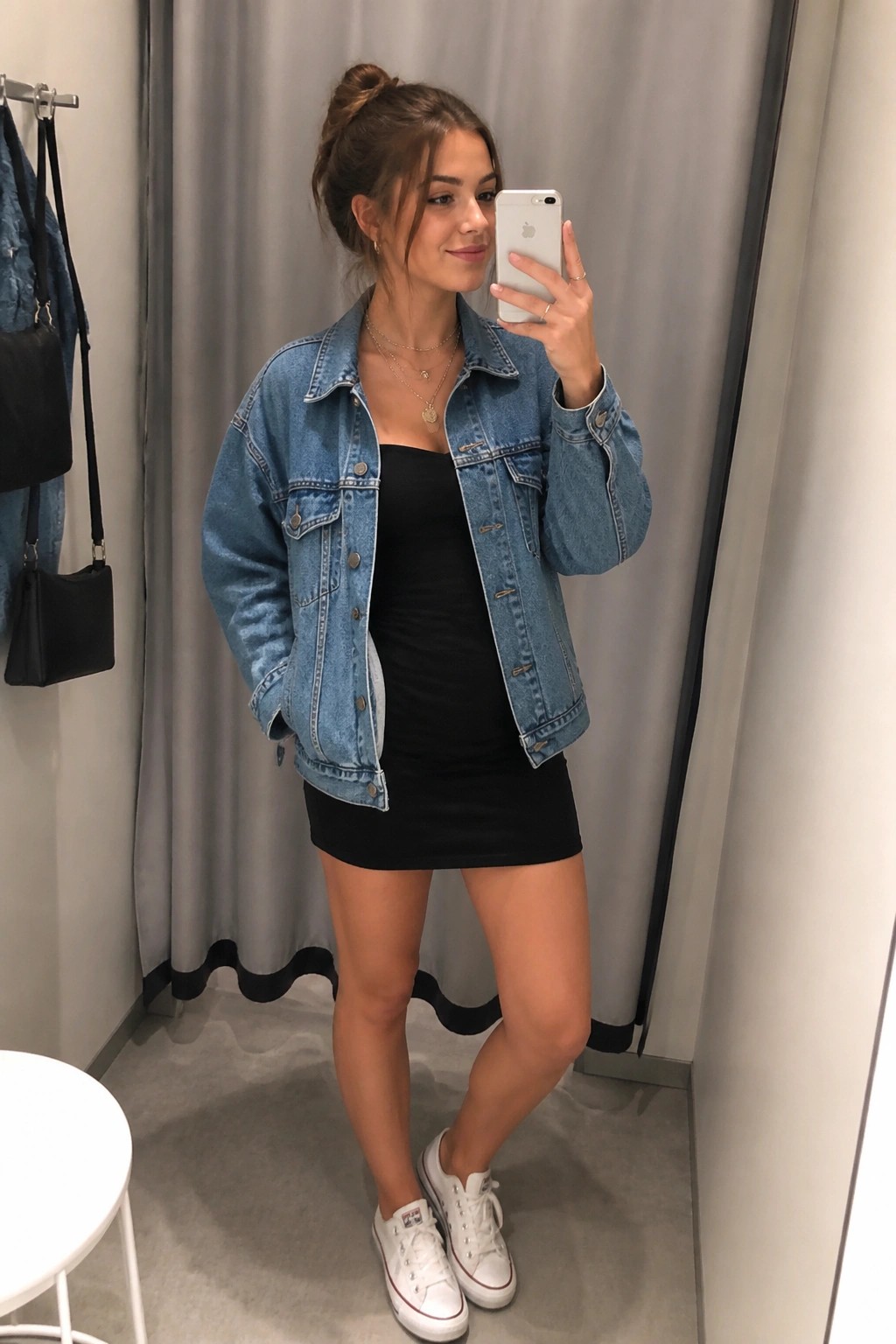 Woman with hair in bun taking mirror selfie in denim jacket and black mini dress