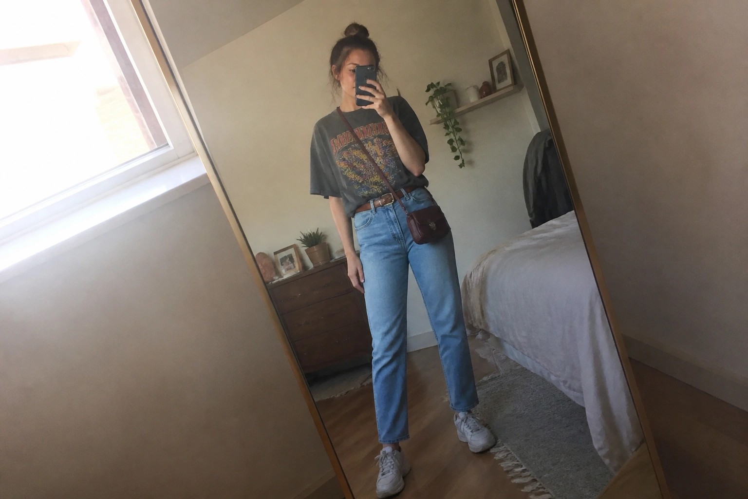 Woman in gray graphic tee and blue jeans taking mirror selfie in bedroom