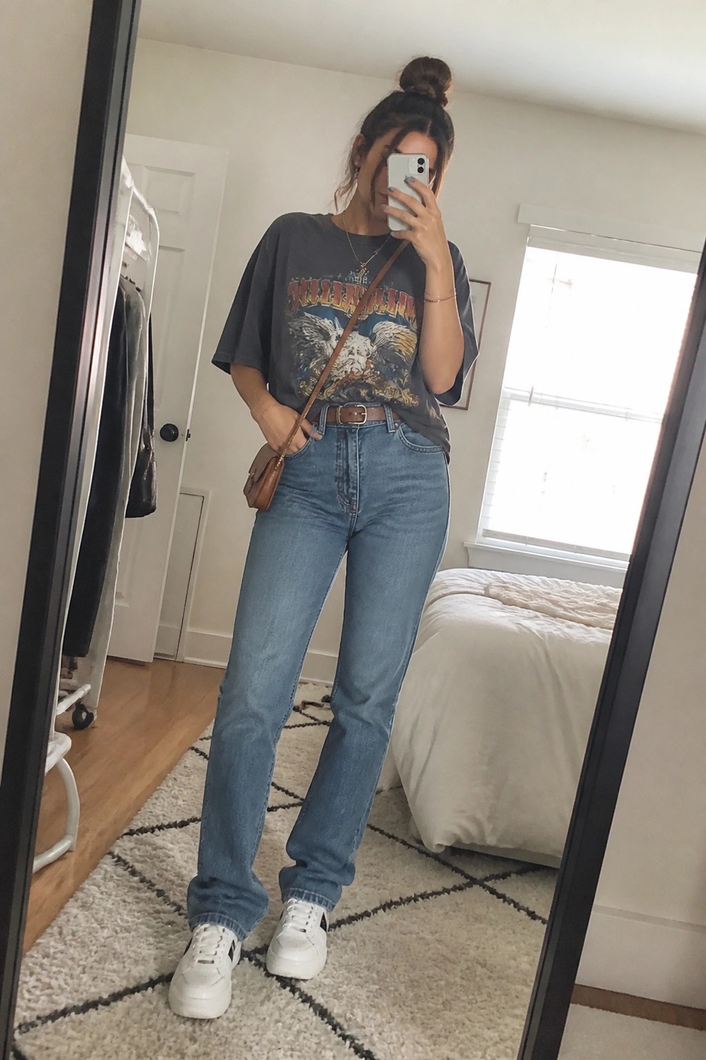 Woman in gray graphic tee and blue jeans taking mirror selfie in bedroom