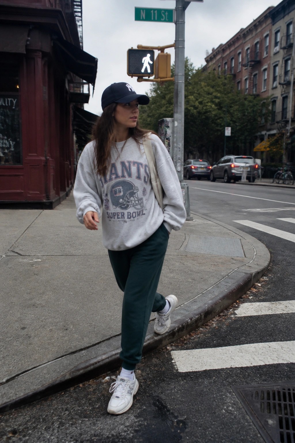 Woman in grey oversized Giants Super Bowl sweatshirt, dark green joggers, white New Balance sneakers, and navy NY cap standing on a city sidewalk.