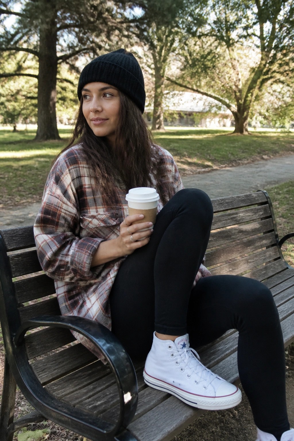 Woman in black beanie and plaid shirt holds coffee on park bench