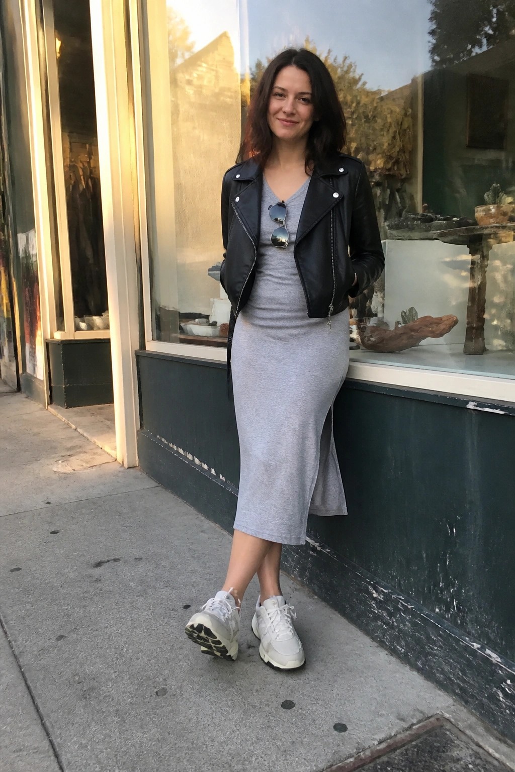Woman wearing a grey midi dress under a black cropped leather jacket with white sneakers and sunglasses tucked into the neckline standing on a sidewalk.