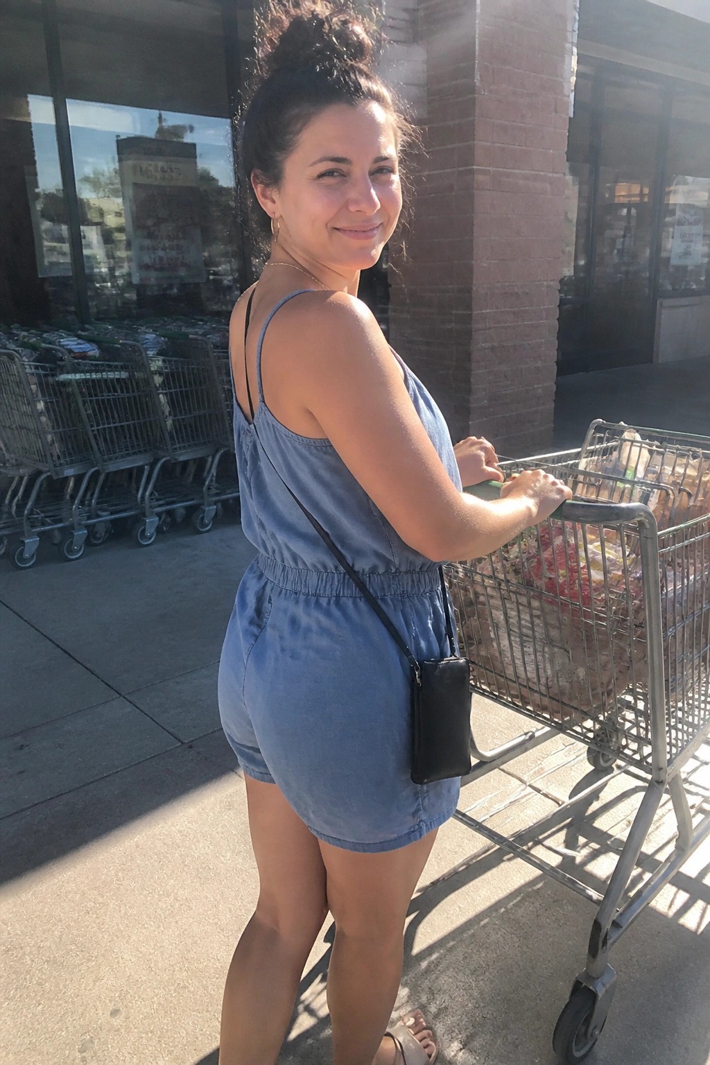 Woman in a light blue denim romper with thin straps and elastic waist paired with a small black crossbody bag standing beside a shopping cart.