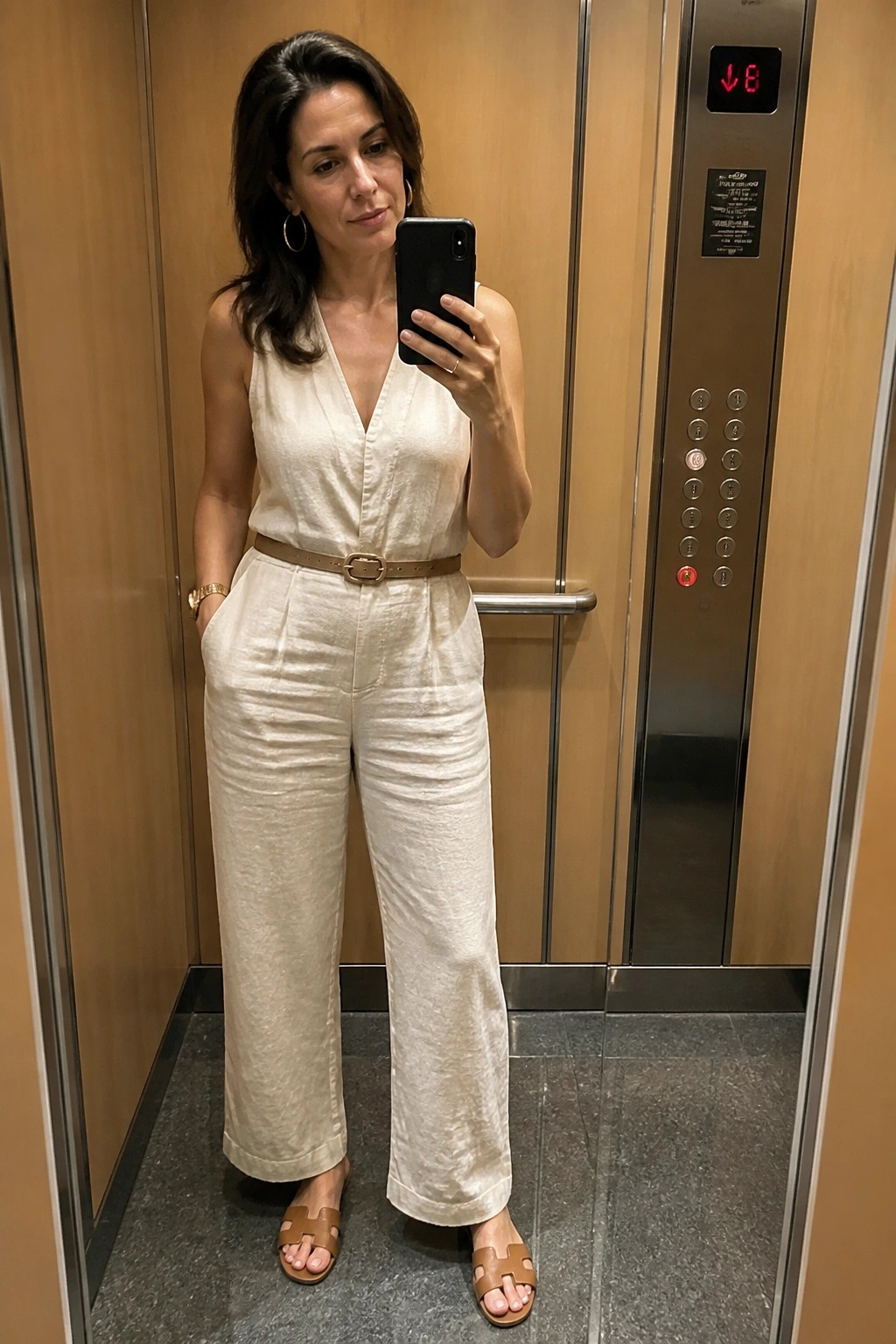 Woman in cream linen jumpsuit taking mirror selfie in elevator