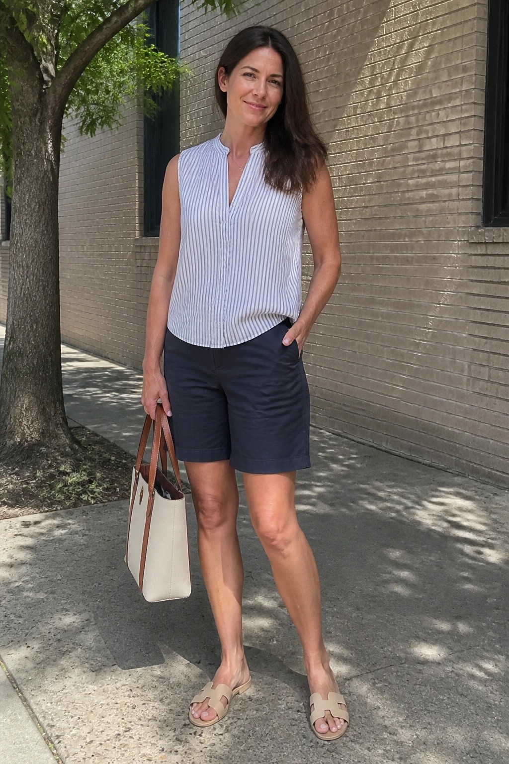 Woman in striped sleeveless top and navy shorts holding tote bag on sidewalk