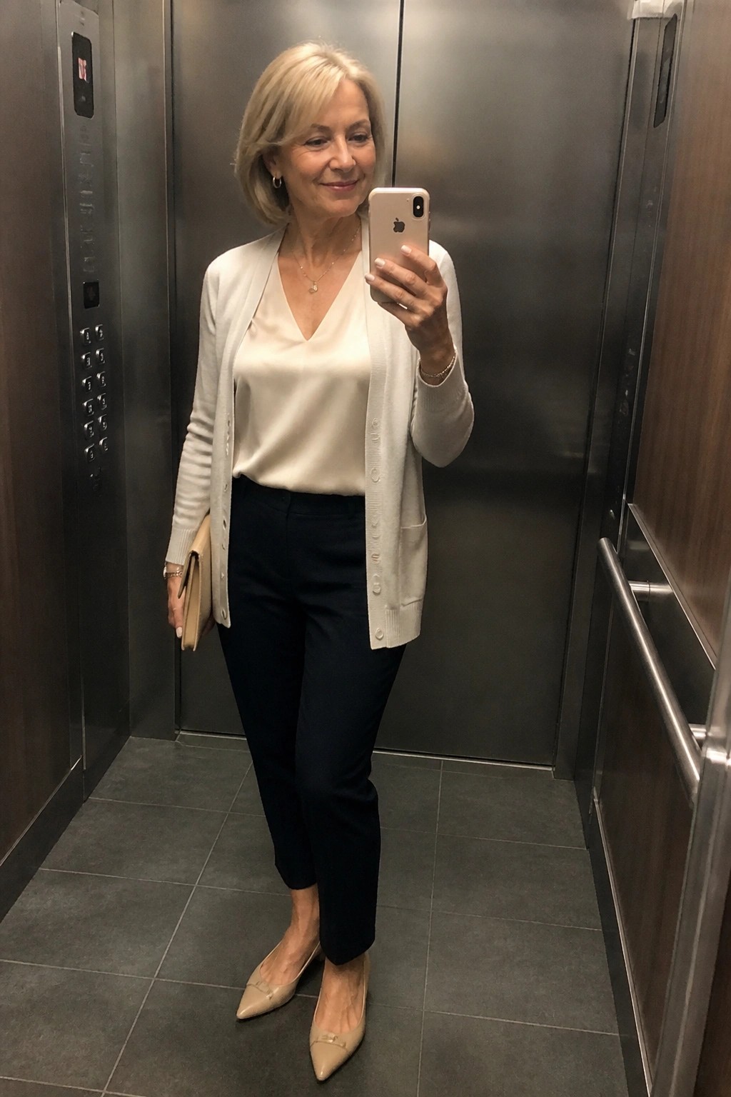 Woman in cream cardigan taking mirror selfie inside elevator with phone