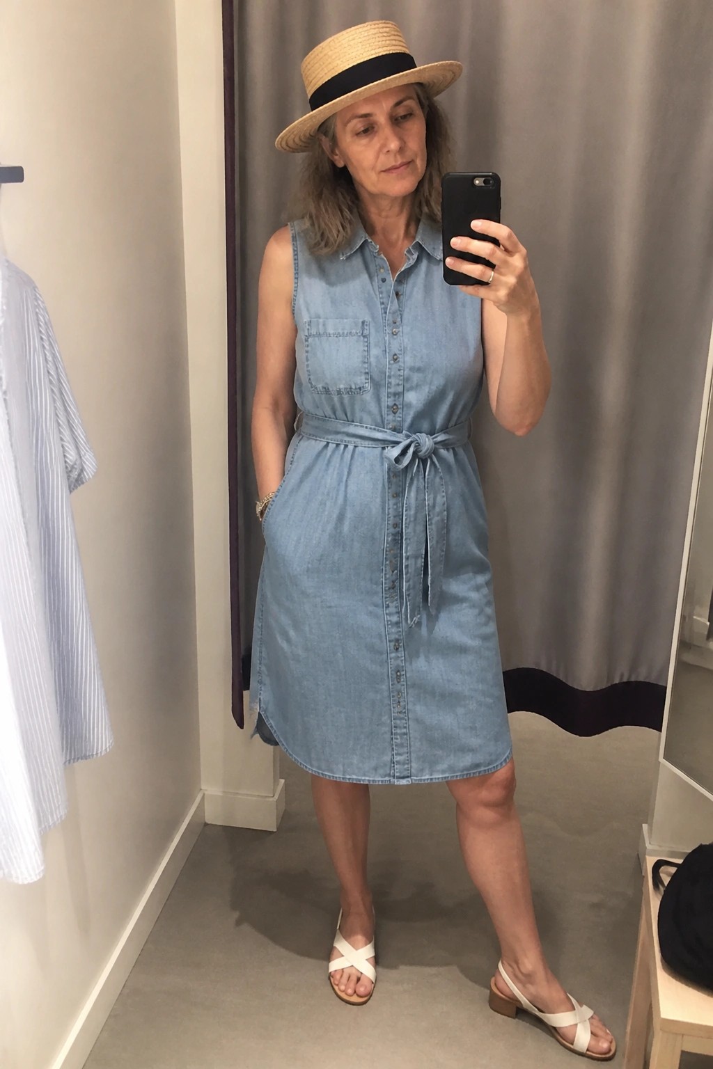 A woman wearing a light blue sleeveless denim shirt dress with a tied belt, a straw hat with black band, and white cross-strap sandals, taking a mirror selfie.