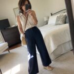 Woman in beige sleeveless shirt and navy pants taking bedroom mirror selfie