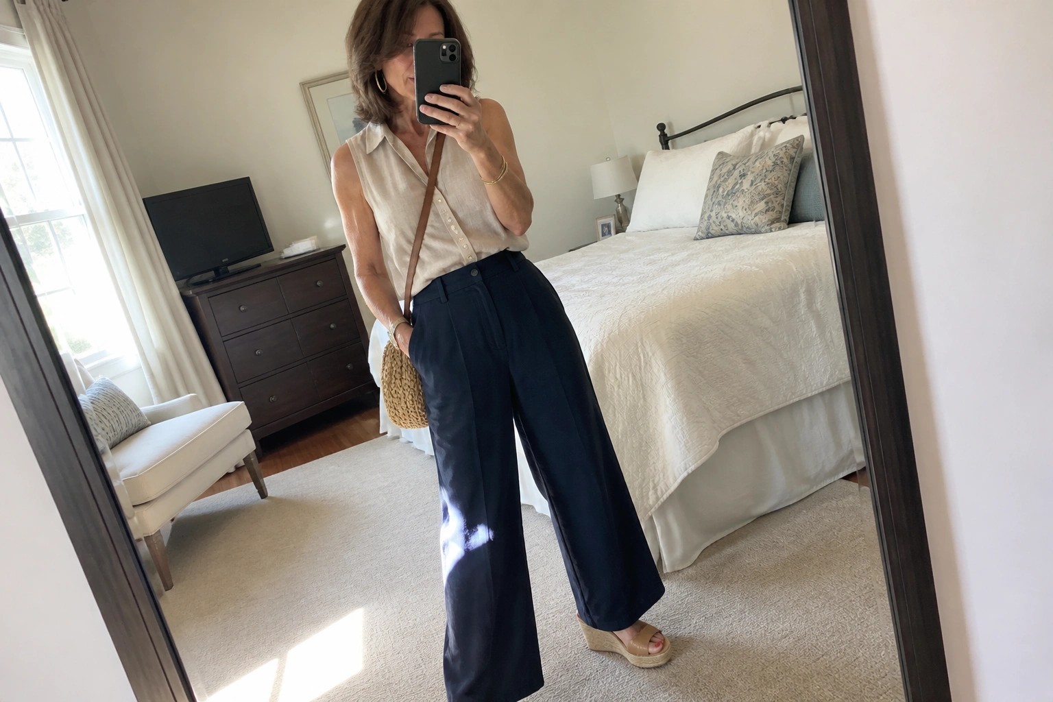 Woman in beige sleeveless shirt and navy pants taking bedroom mirror selfie