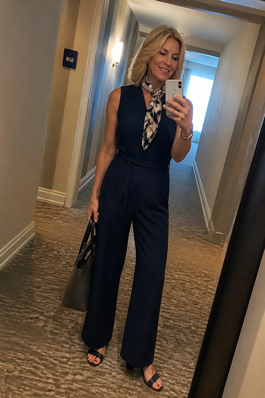 Woman in navy jumpsuit with scarf taking mirror selfie in hotel hallway holding bag