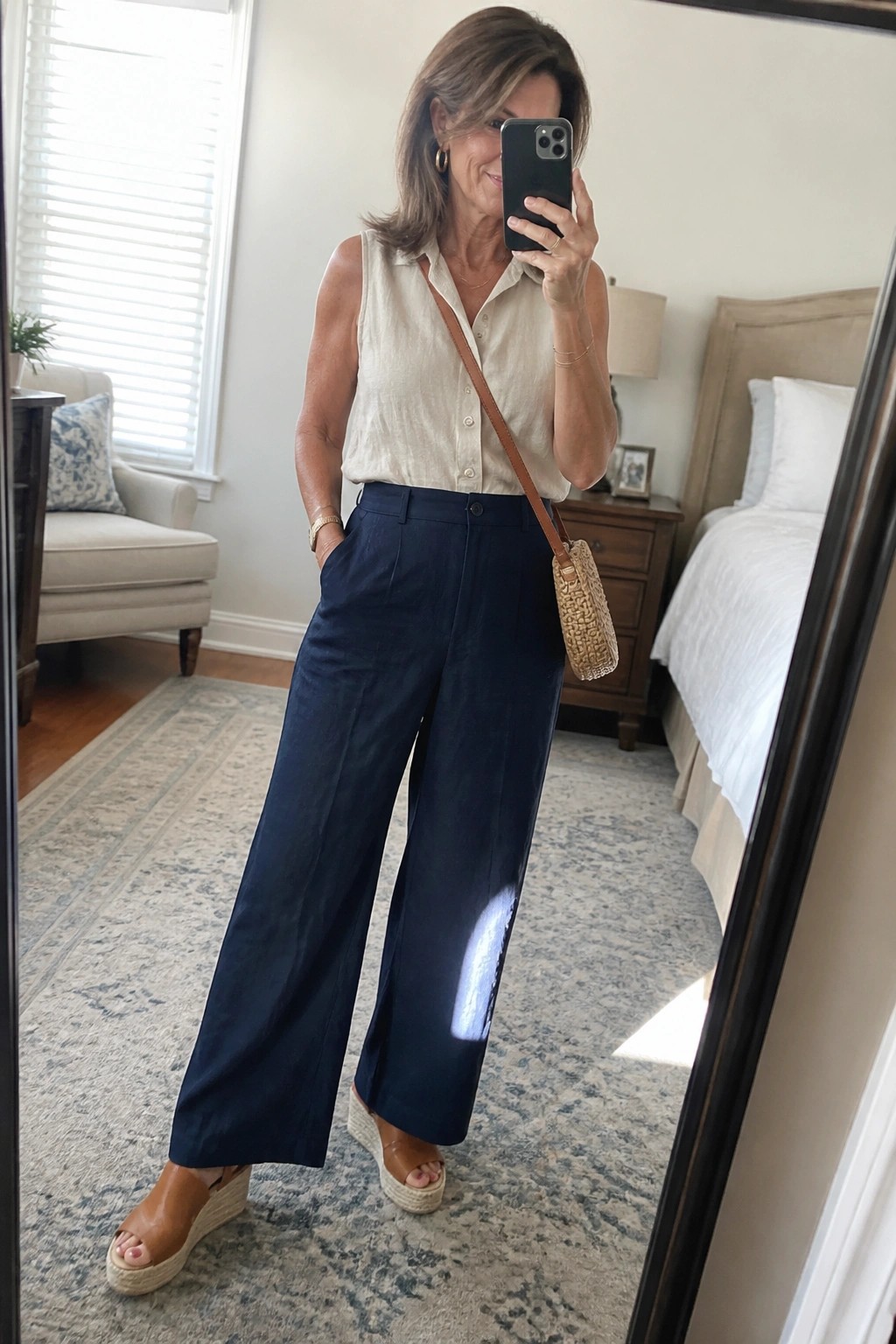 Woman in beige sleeveless shirt and navy pants taking bedroom mirror selfie