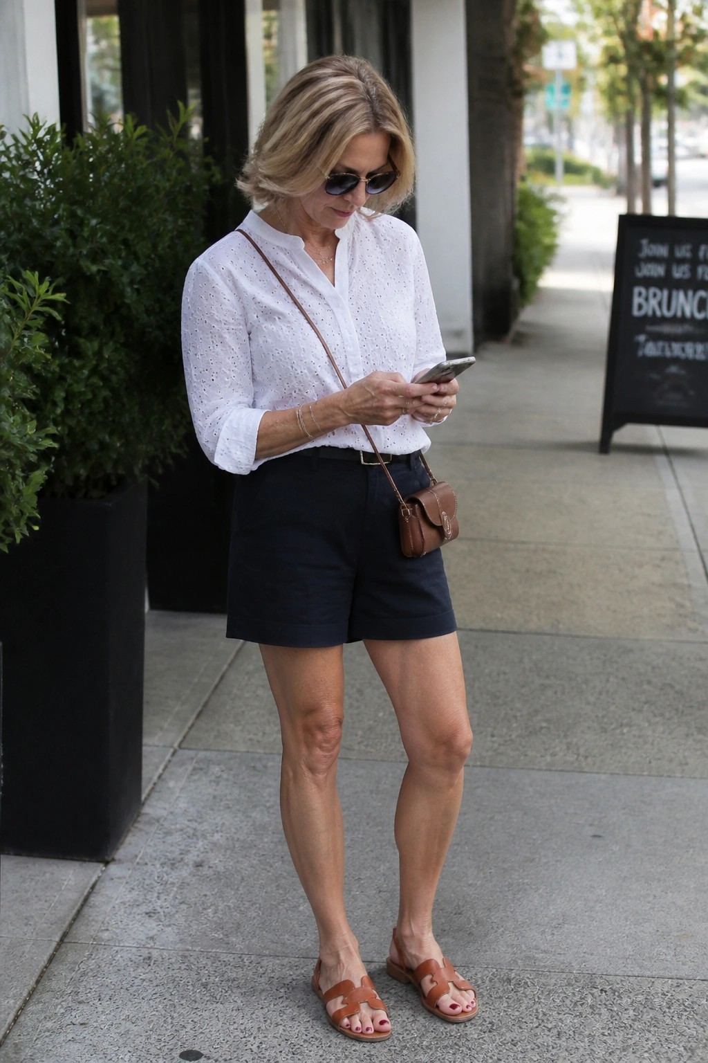 Woman in white eyelet blouse and navy shorts checks phone on sidewalk
