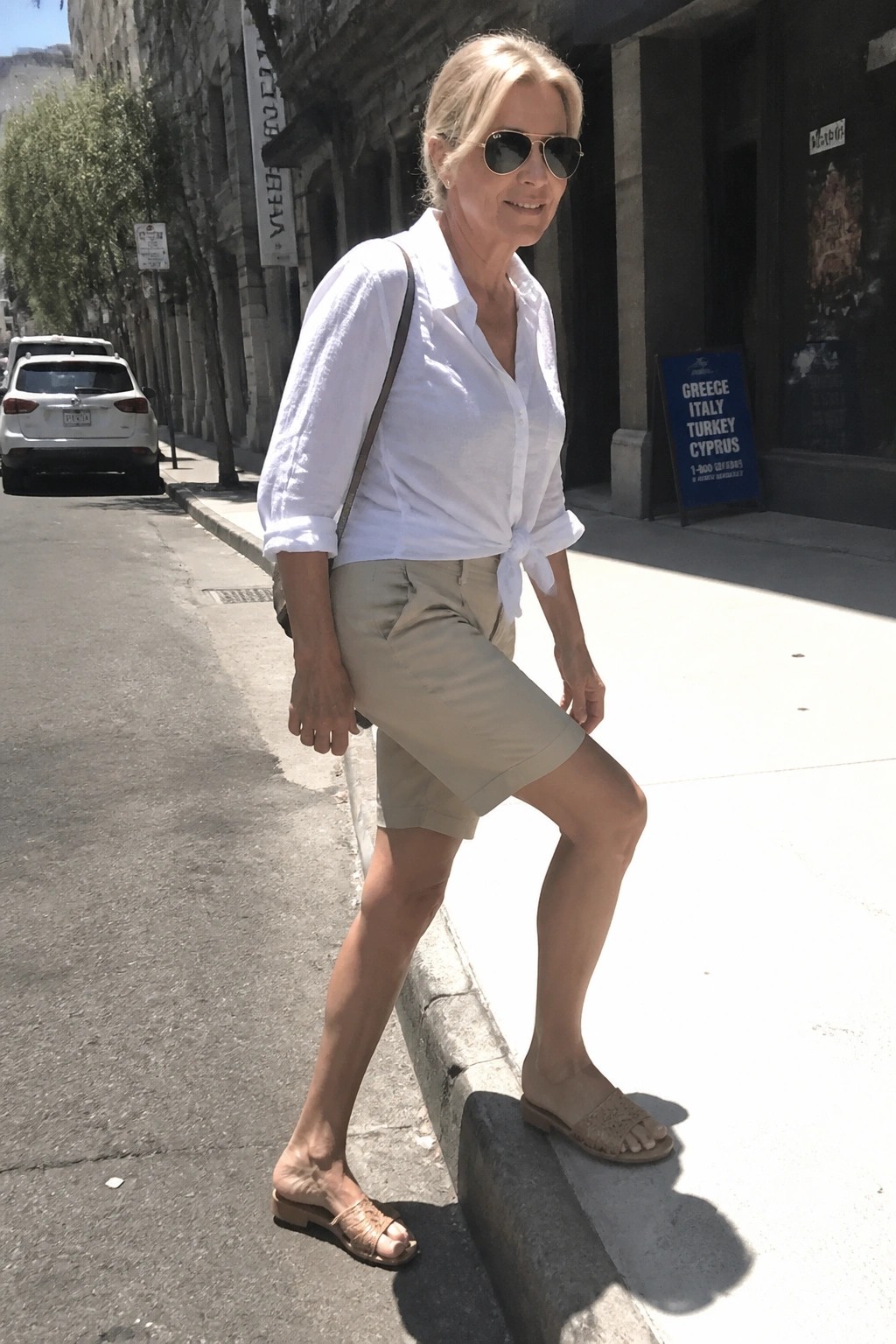 Blonde woman in white tied shirt and beige shorts on sunny sidewalk