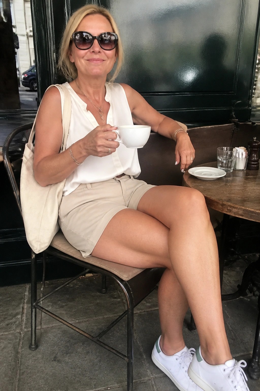 Woman over 50 in white sleeveless blouse tucked into beige shorts with white sneakers, layered necklaces, bracelets, beige tote bag over shoulder, holding white coffee cup while seated outdoors.