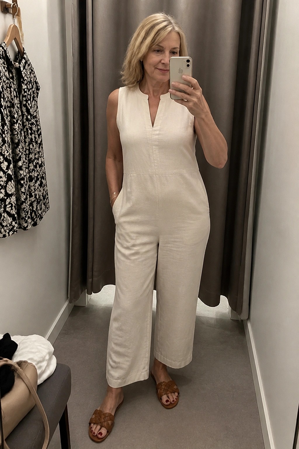 Woman over 50 wearing a cream sleeveless linen jumpsuit with wide legs and brown woven leather slides, taking a mirror selfie in a fitting room.