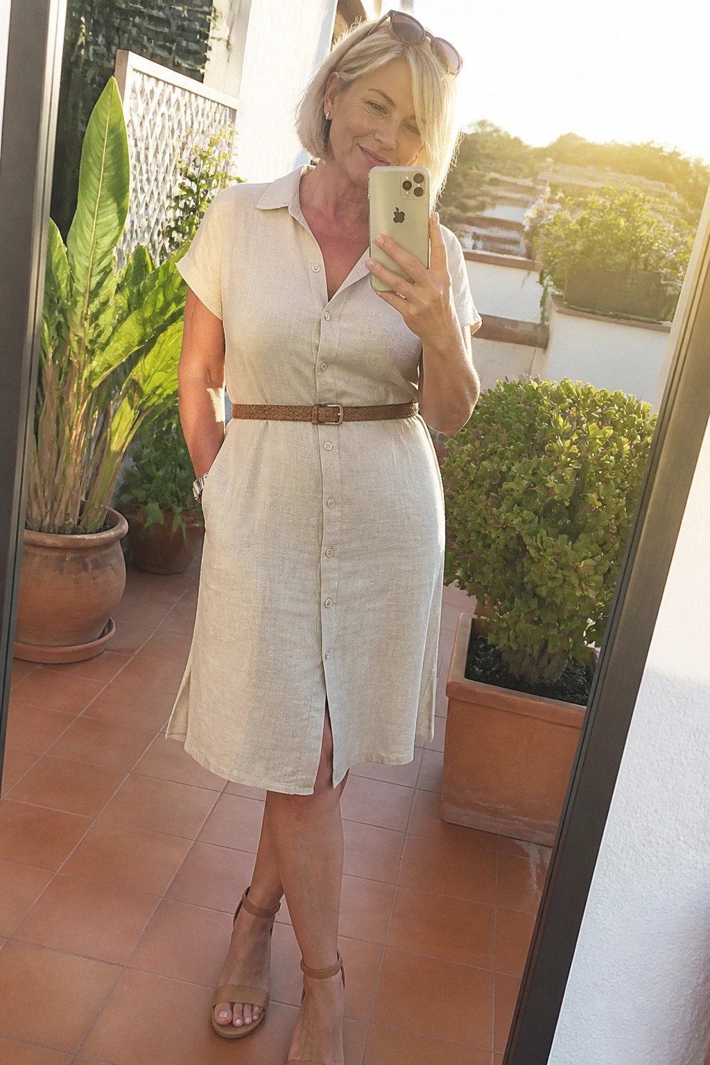 Woman in beige linen shirt dress taking selfie on sunny patio with plants