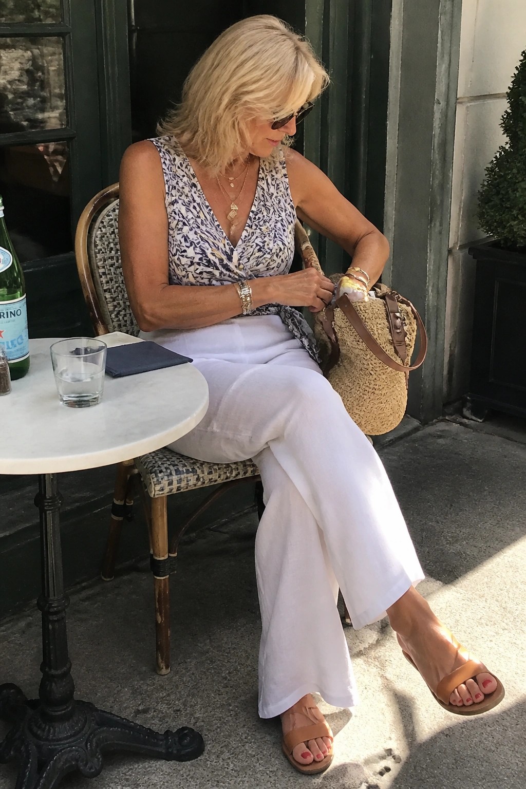 Blonde woman in patterned top and white pants holding straw bag at cafe table