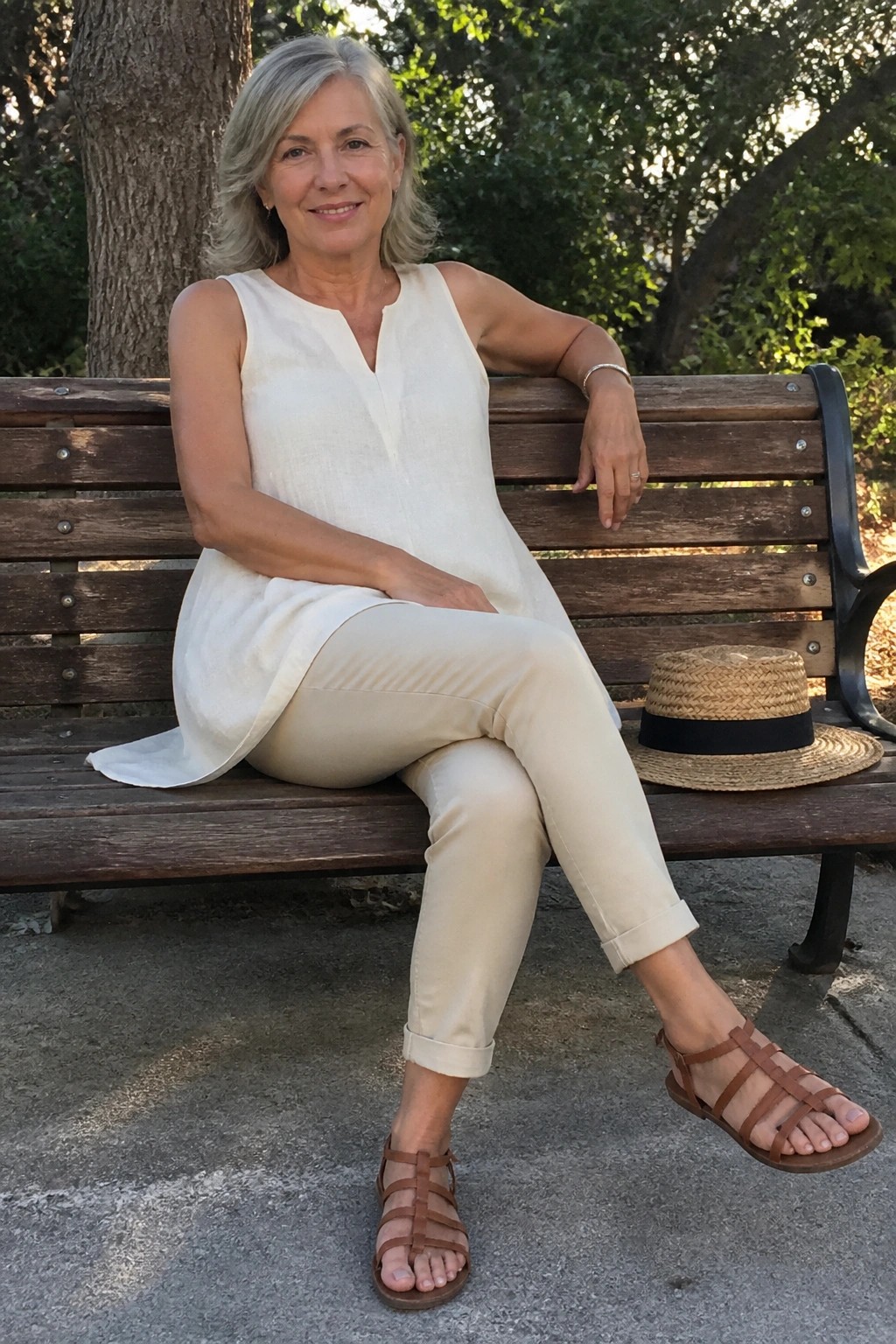 Gray-haired woman in white top and beige pants sits on bench with straw hat