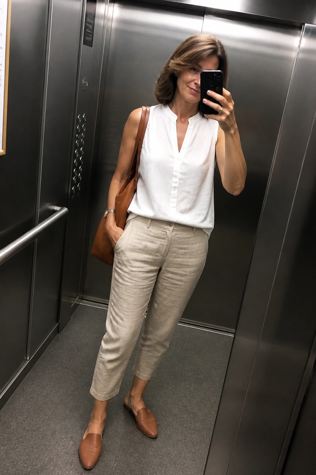 Woman in white top and beige pants taking selfie in elevator with brown bag