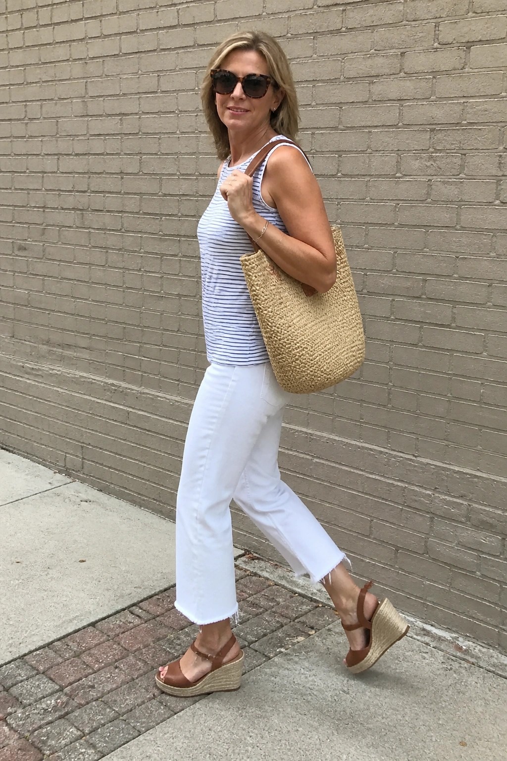 Woman over 50 in striped sleeveless tank, white cropped jeans with frayed hems, brown wedge sandals and woven straw tote bag standing against brick wall.