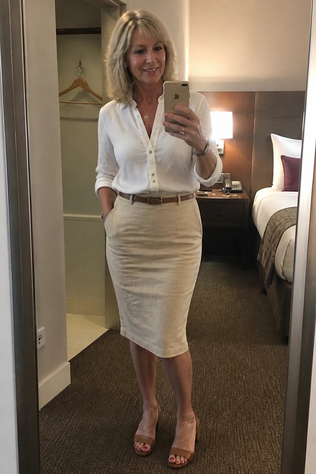 Woman over 50 wearing a white linen button-up shirt tucked into a beige linen pencil skirt with brown belt and matching block-heel sandals while taking a mirror selfie.