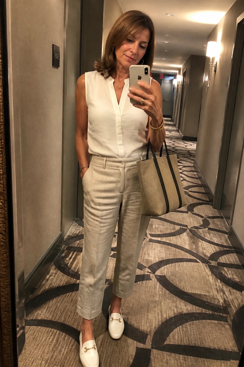 Woman over 50 in white sleeveless linen blouse tucked into beige linen trousers with white horsebit loafers and beige black striped tote bag standing in hallway taking mirror selfie.