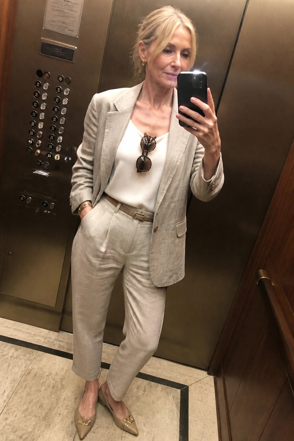 Blonde woman in beige linen pantsuit taking mirror selfie in elevator