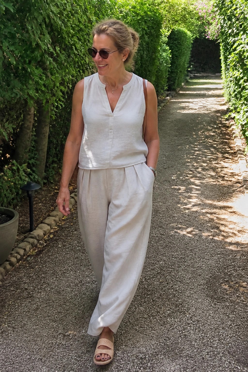 Woman in white linen top and pants walking on sunny garden path between hedges.