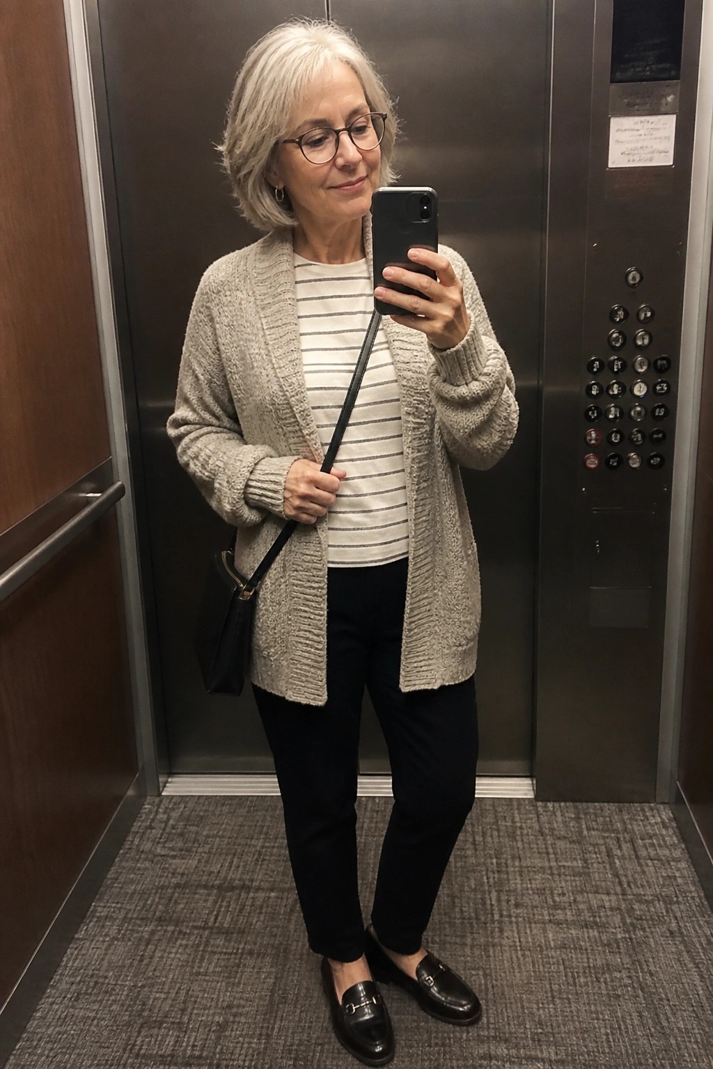 Older woman wearing a light gray chunky knit cardigan over a white striped long sleeve top with black pants and black loafers while holding a phone in an elevator mirror selfie