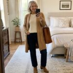 Woman in tan cardigan and jeans taking mirror selfie in bedroom with leather bag
