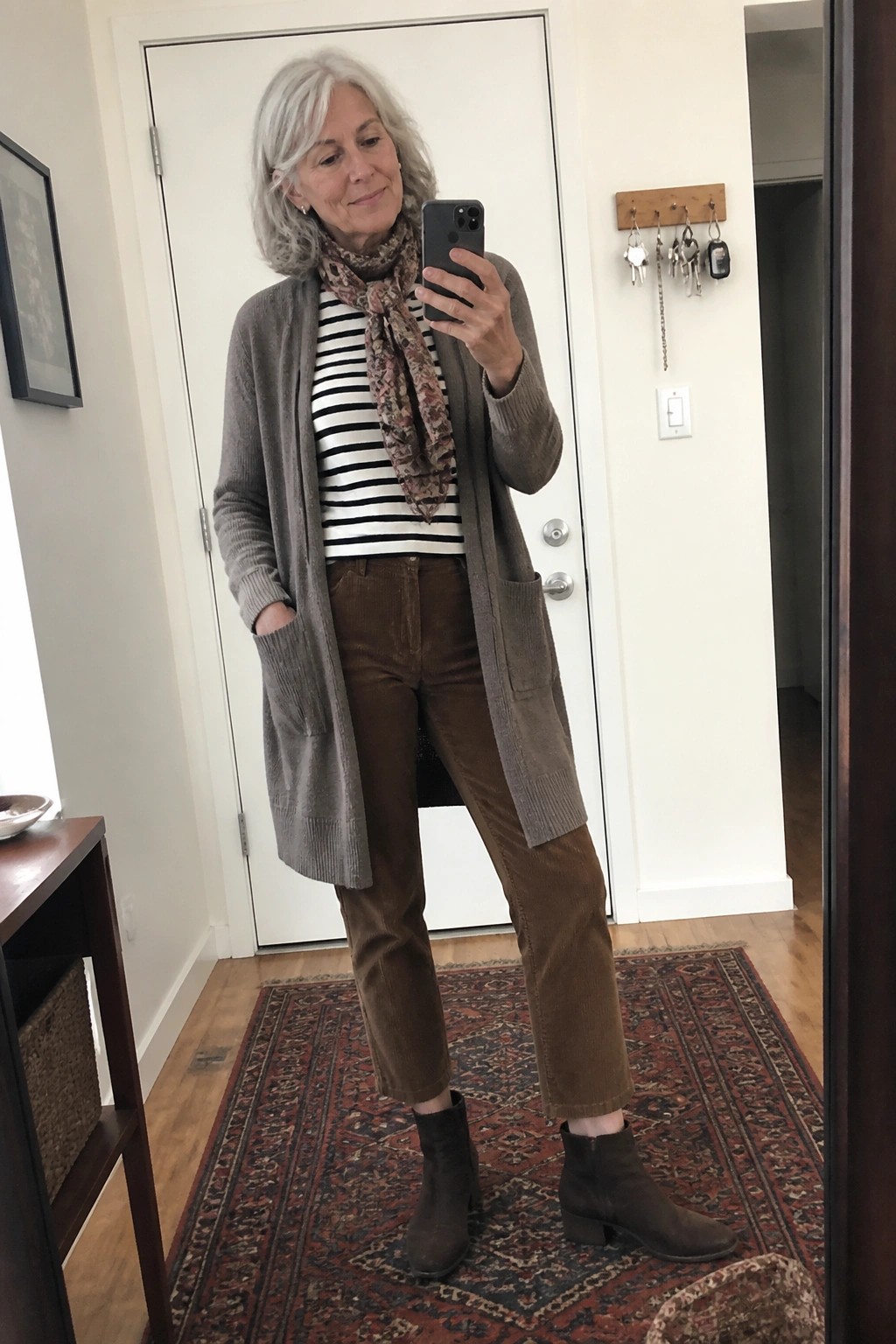 Woman with gray hair taking mirror selfie in gray cardigan and brown corduroy pants