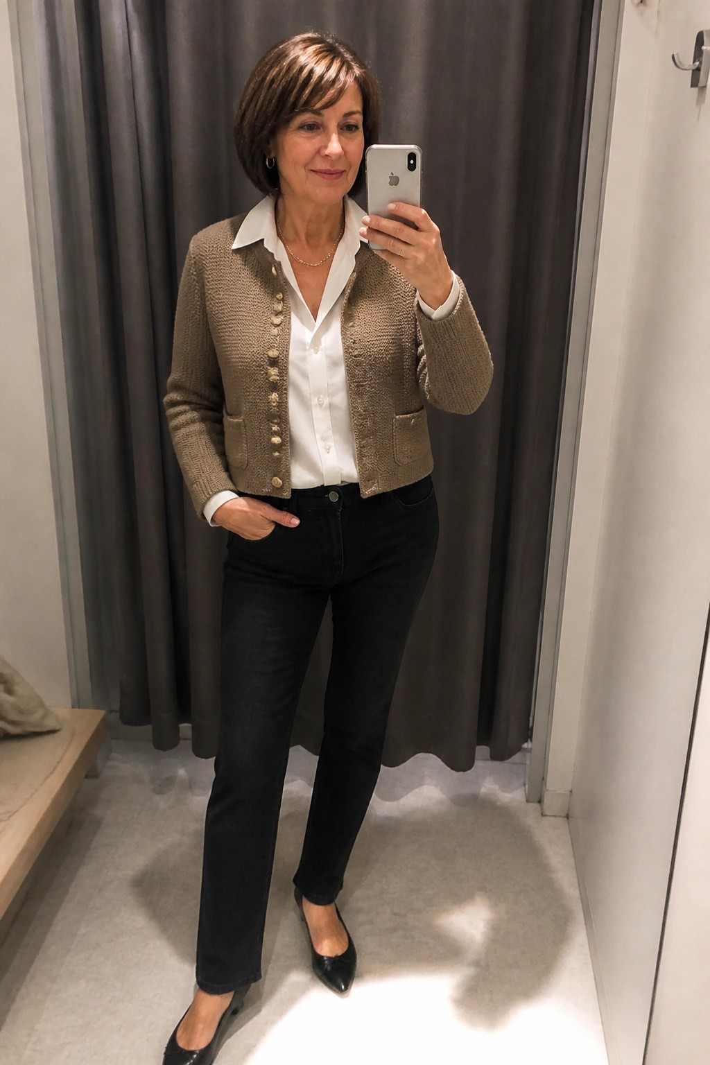 Woman in brown knit cardigan and black jeans taking mirror selfie in fitting room
