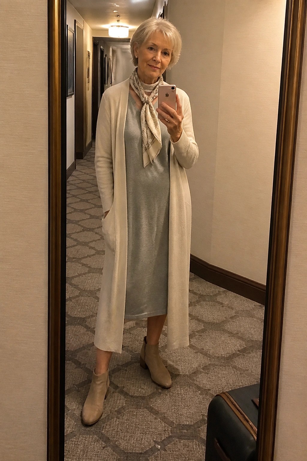 Woman in light gray dress and cream cardigan taking mirror selfie in hallway