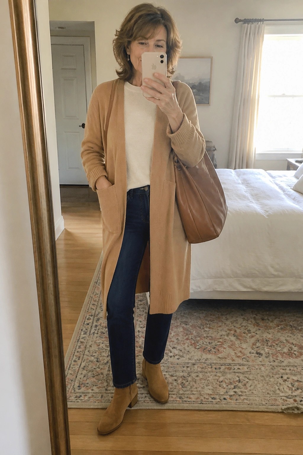 Woman in tan cardigan and jeans taking mirror selfie in bedroom with leather bag