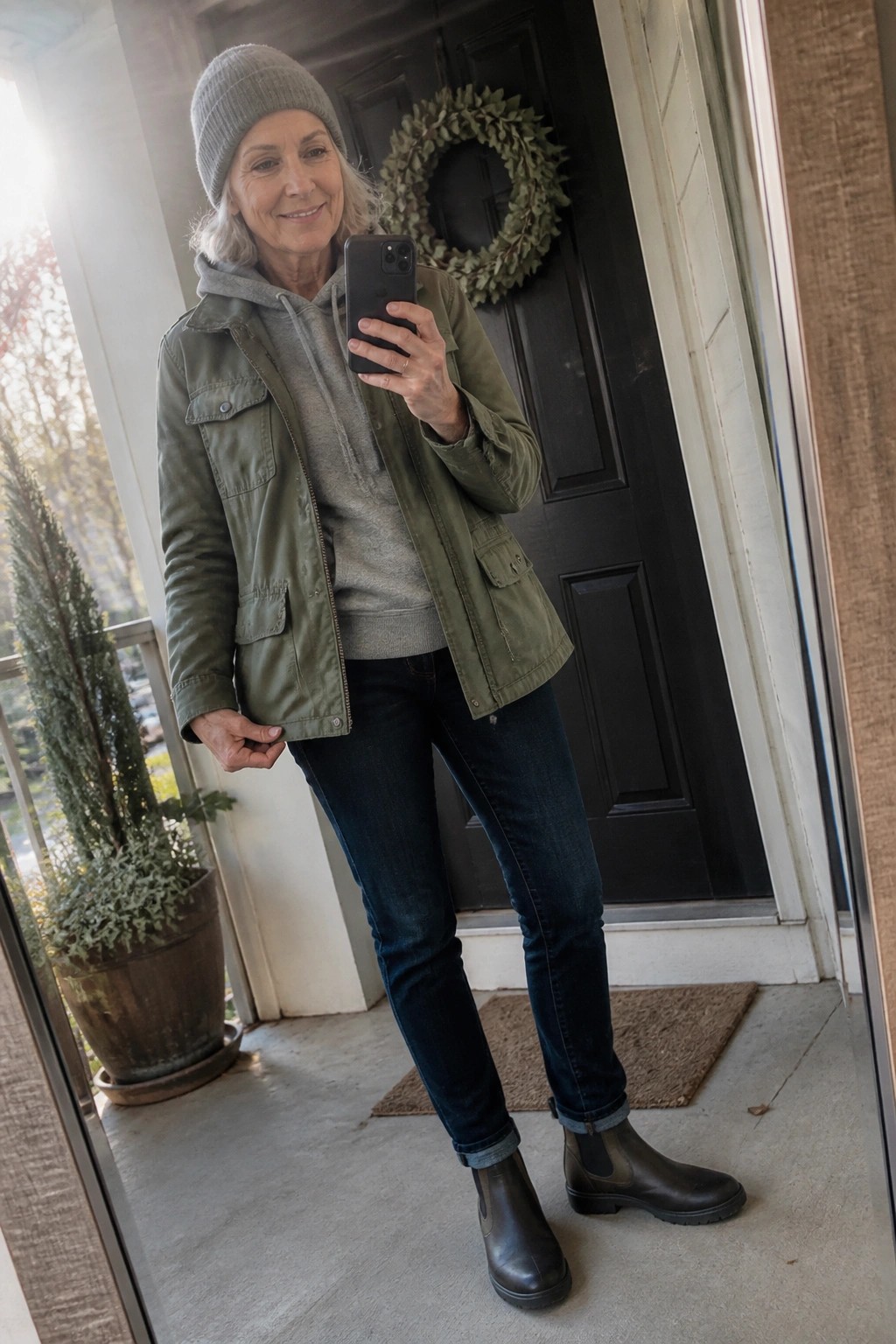Older woman in green jacket and beanie taking selfie on porch with phone