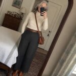 Woman with short gray hair wearing a white turtleneck sweater, dark gray wide leg pants with brown belt, brown leather crossbody bag, and brown ankle boots while taking a mirror selfie in a bedroom.