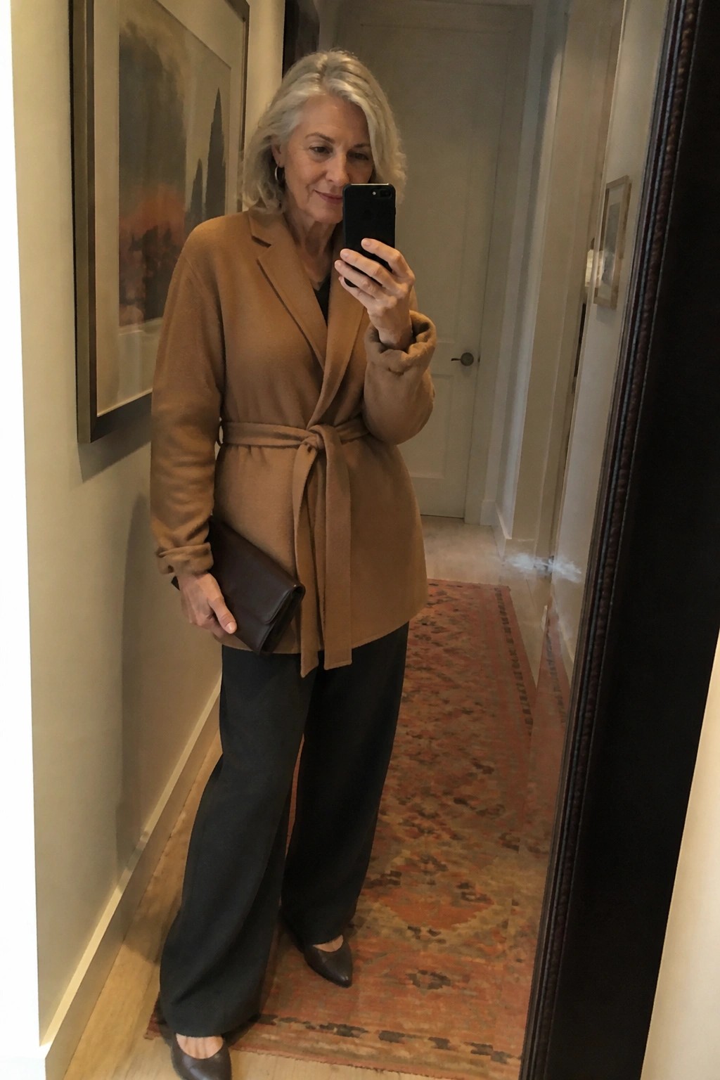 Gray-haired woman in brown belted coat takes mirror selfie in hallway.