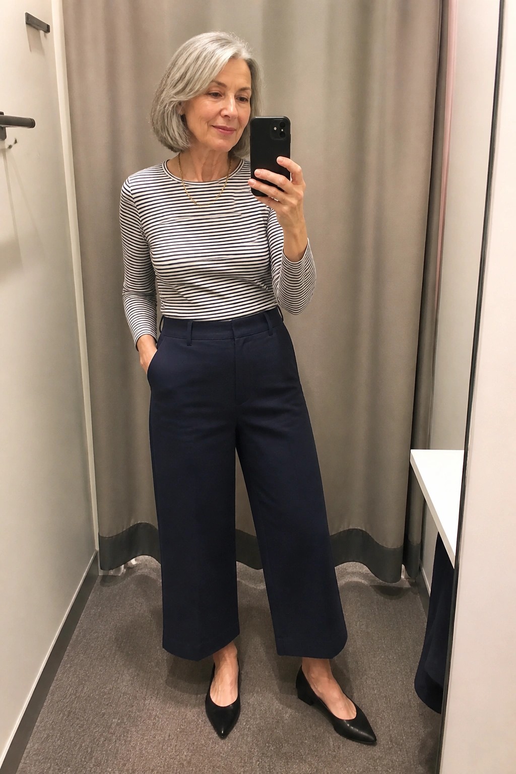 Gray-haired woman in striped top and navy wide-leg pants taking mirror selfie.