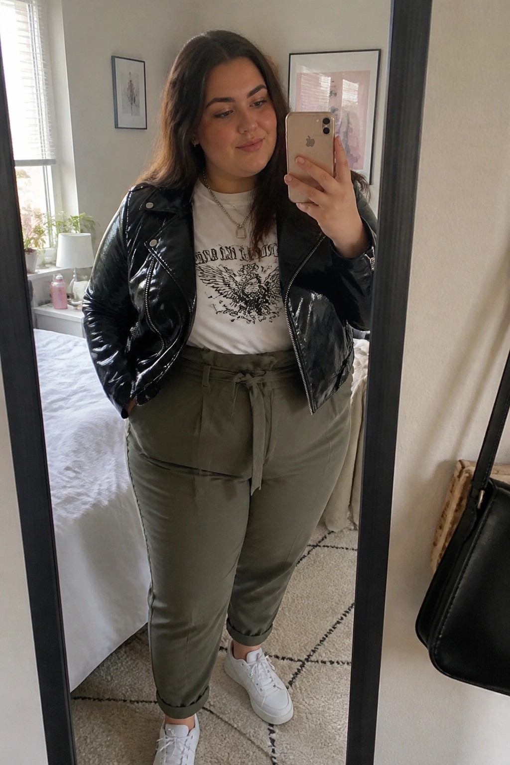 Woman in glossy black jacket, graphic tee, and olive pants taking mirror selfie