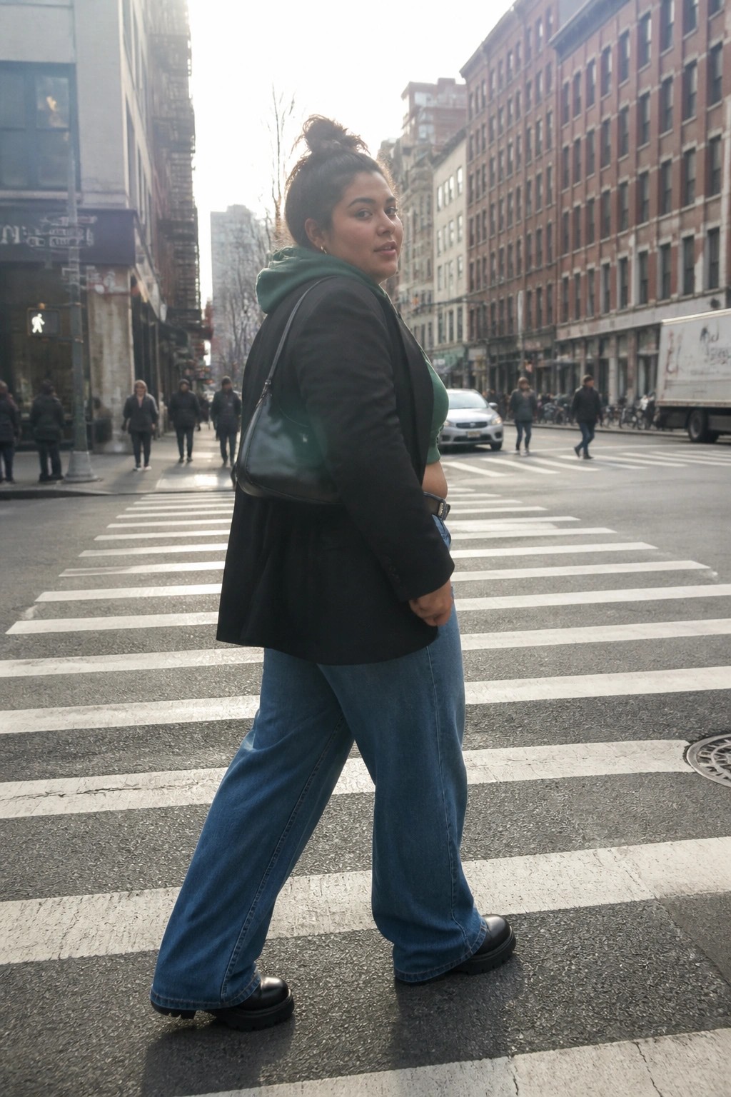 Woman in black blazer and blue jeans crossing sunny city crosswalk, looking back.